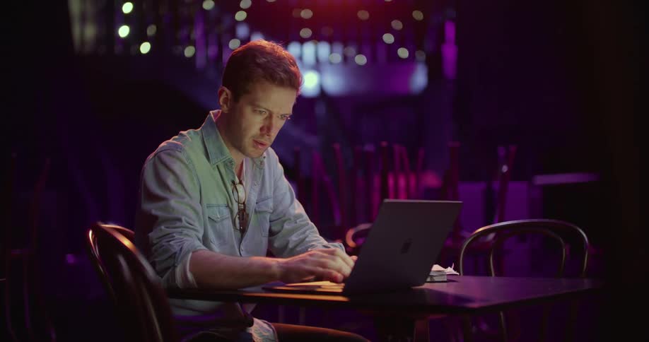 A man in a nightclub sitting on a chair at a table using a laptop