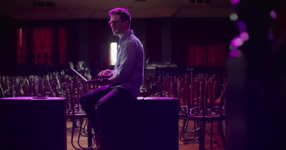 A man sits against a table working on a laptop with lots of tables and chairs surrounding him