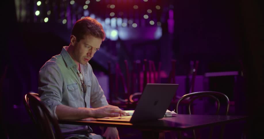A man flips through a book while using a laptop at a table in a dimly lit closed restaurant