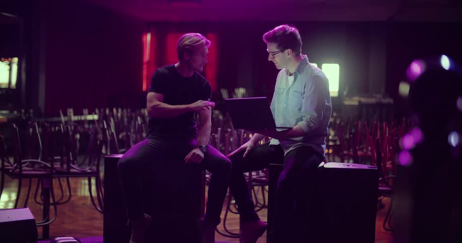 Two men sit on a stage of a nightclub while talking as one holds onto a laptop computer