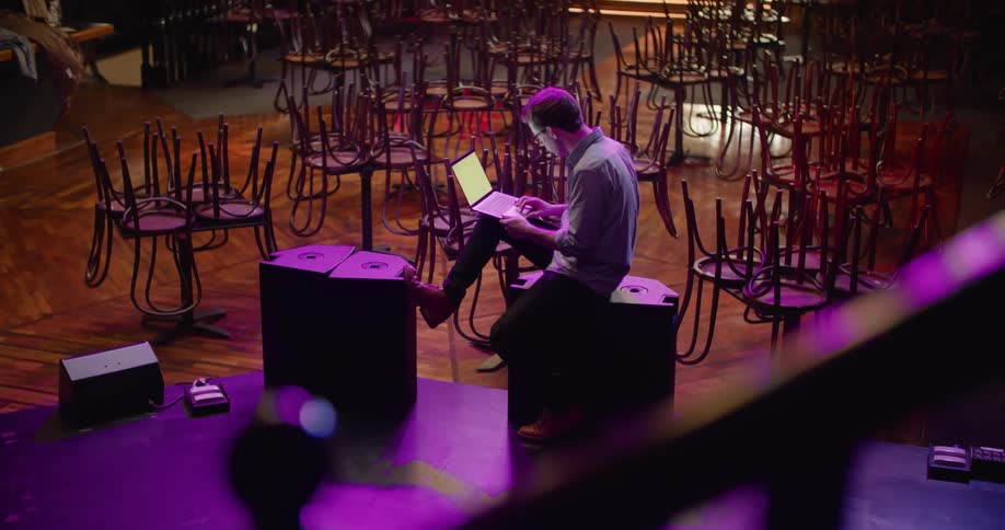 Man with glasses and shirt uses laptop on the bar stage against upside down chairs on tables in the bar