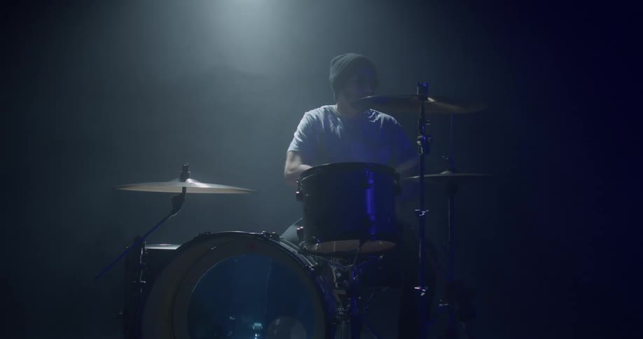 Drummer wearing knit hat plays drums on smoky stage under spotlight