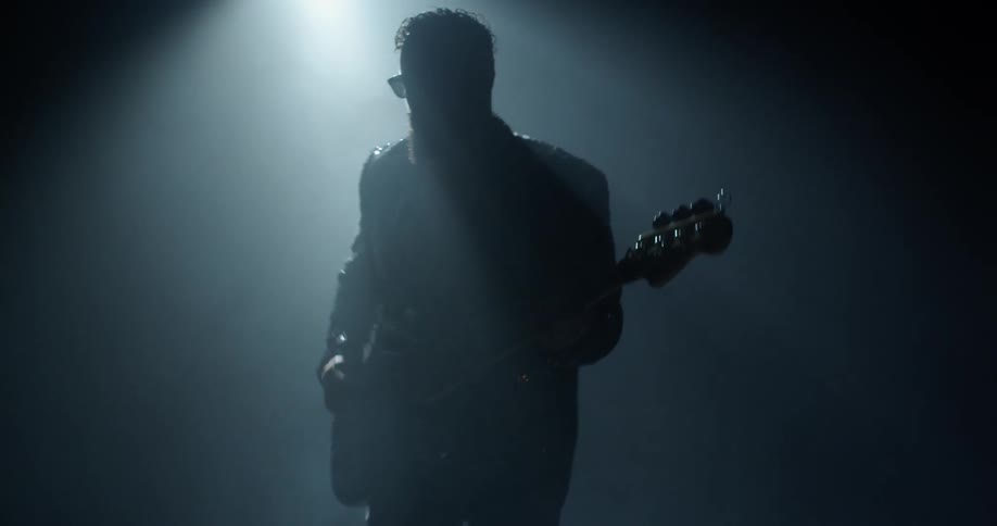 Guitarist with beard wears glasses while playing guitar on foggy stage under bright light