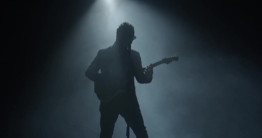 Man wearing glasses sings into a microphone as he plays a guitar