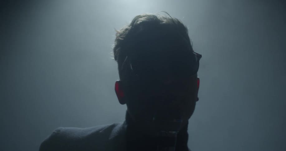 A man wearing sunglasses indoors sings emphatically into a microphone
