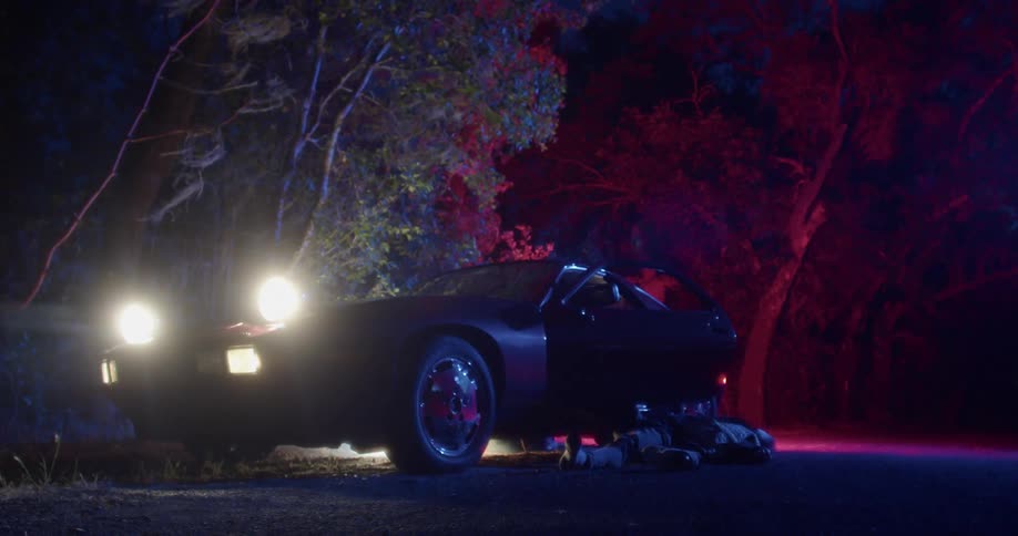 A man sits in the car with a body laying on the floor outside