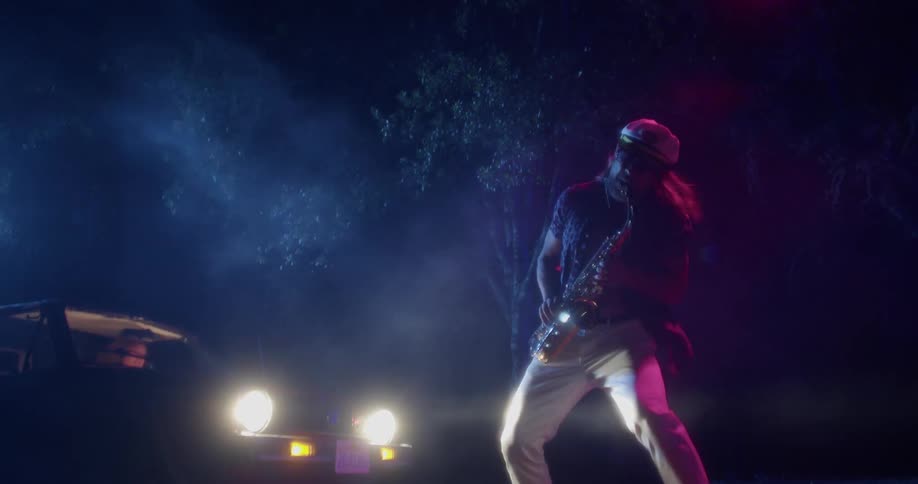 Man wearing a cap plays a saxophone outdoors as a cars headlights shine on him