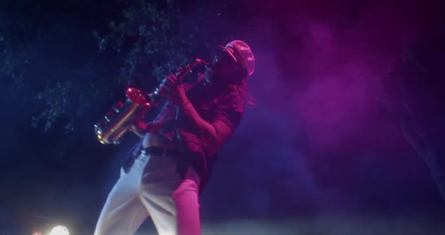 Car headlights shine on a man wearing glasses and playing a saxophone near the trees