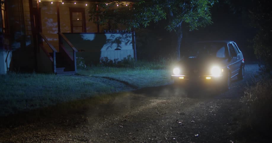 A vehicle with headlights in a driveway next to a house and tree