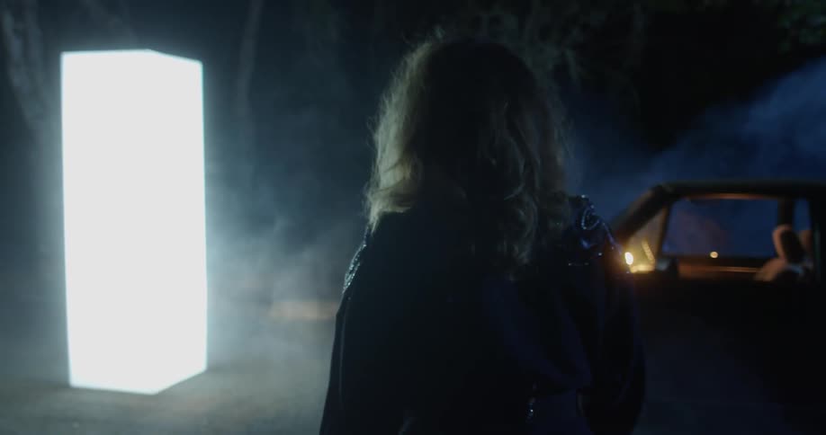 A woman stands next to her vehicle and watches a glowing monolith
