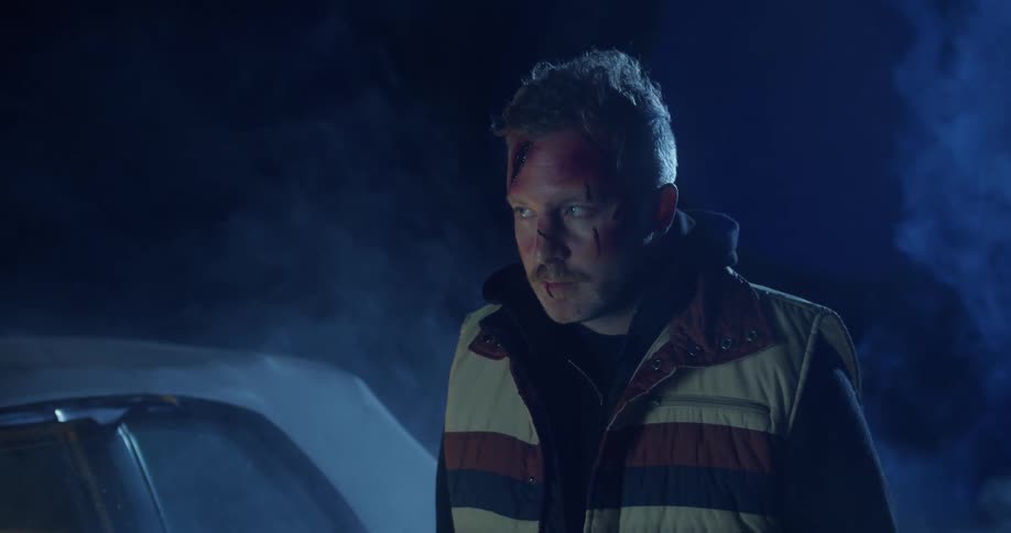 An injured man stands outside of a vehicle at night as fog drifts past
