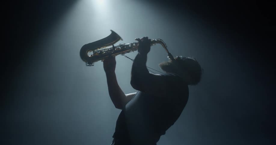 A man in a room playing a saxophone with a spotlight and a wall in the background