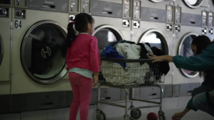 A child kicks a ball to an adult at a laundromat and the ball gets stuck under a laundry cart