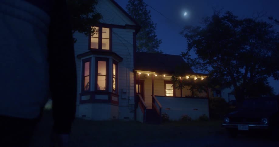 A man walking towards a house with a vehicle in the driveway with the sky clouds and the moon in the background