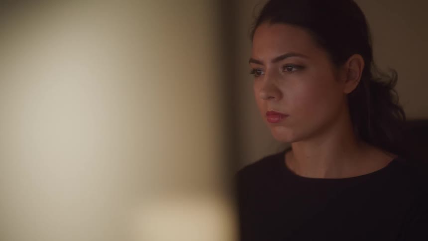 A woman in a room sitting on a chair in front of a monitor with a wall in the background