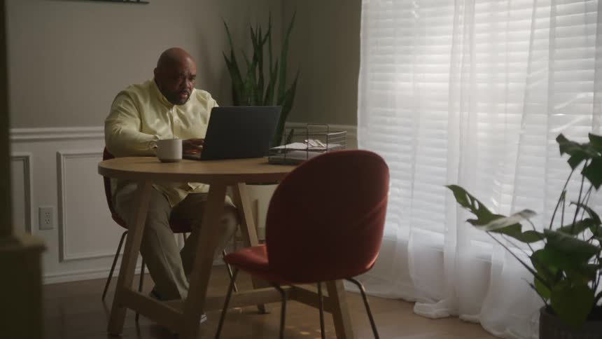 A man in a room sitting on a chair at a table using a laptop