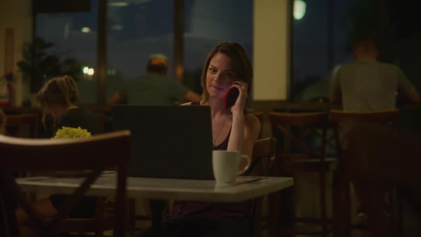 A woman in a coffee shop sitting at a table talking on a smart phone and using a laptop