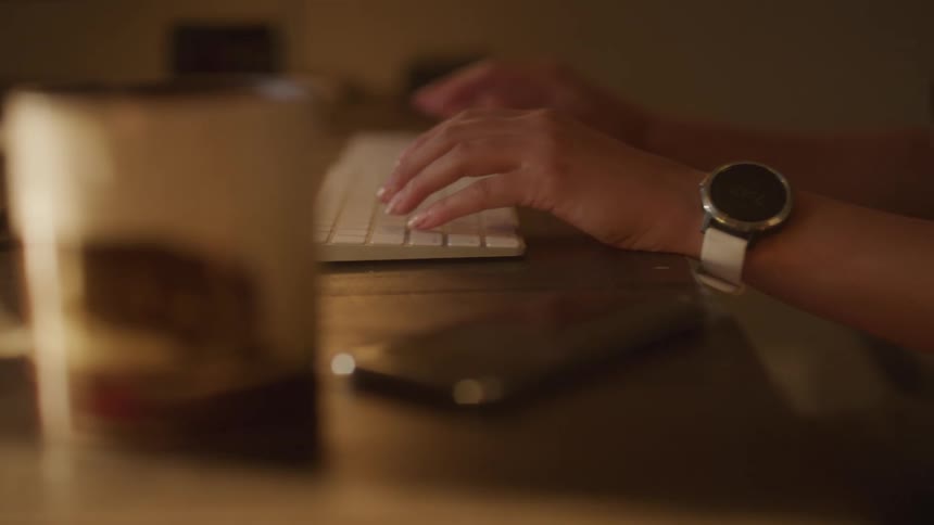 A person sitting at a desk types on a computer keyboard with their smart phone next to it