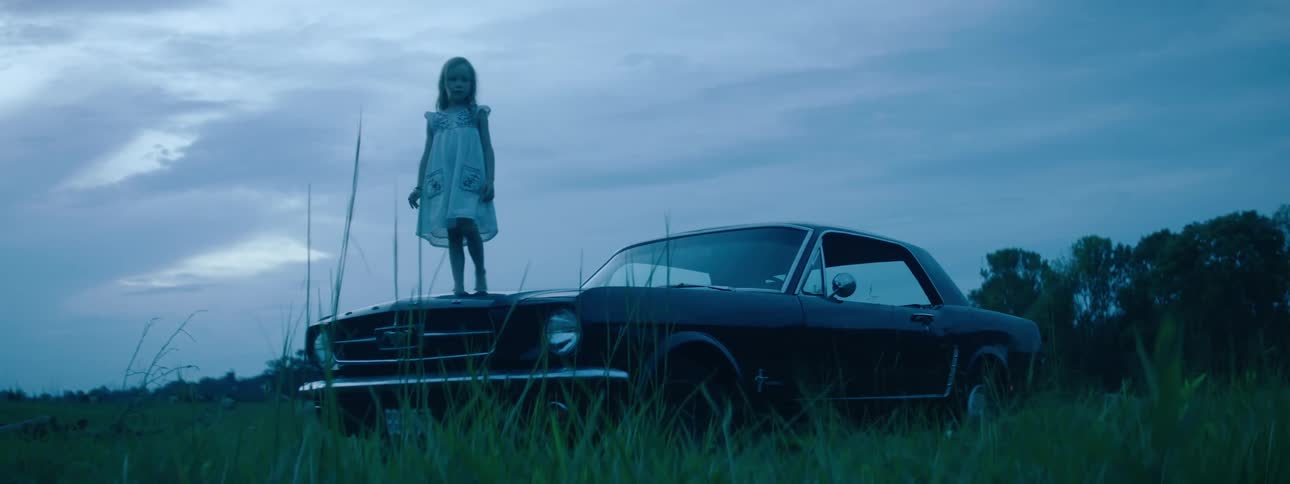 Little girl wearing white dress standing over a muscle car parked on the countryside at morning