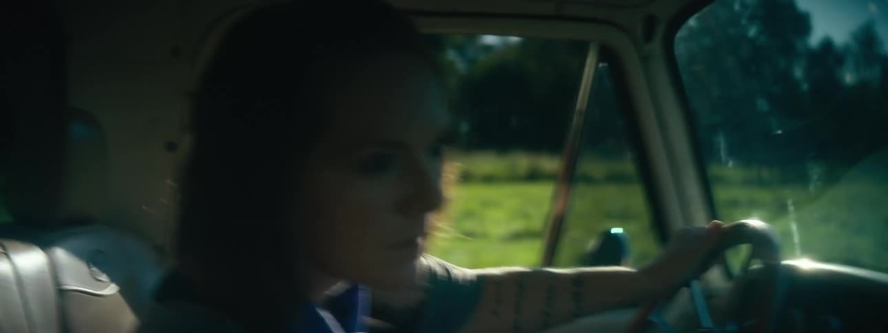 A woman driving a vehicle with a grassy field the sky and clouds in the background