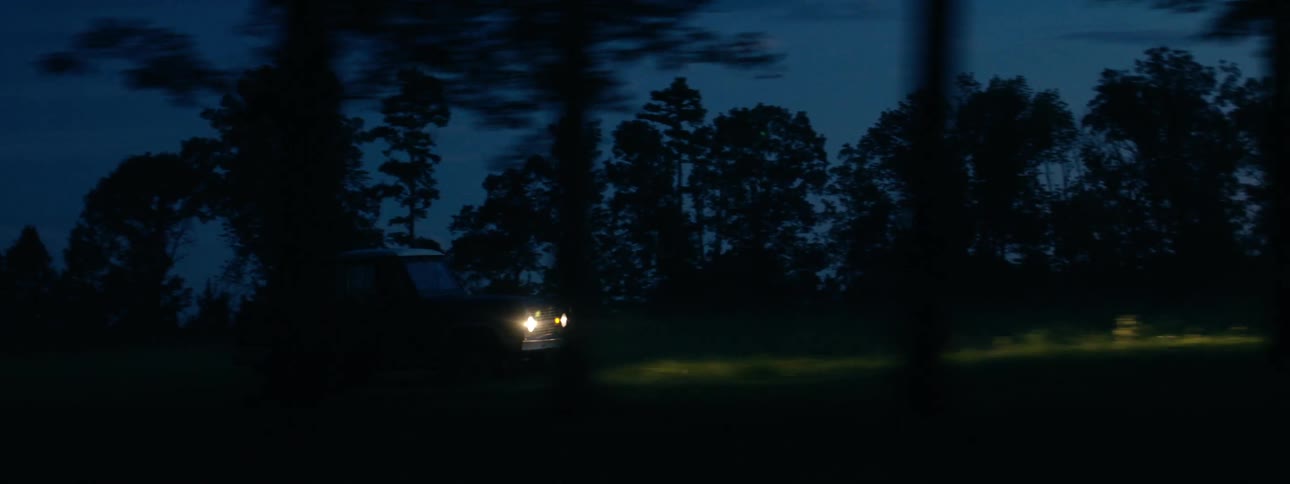 A vehicle with headlights driving on a grassy field with trees the sky and clouds in the background