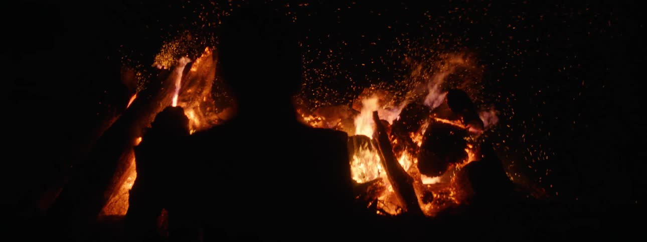 The silhouette of a person in front of a flaming fire with embers glowing at night