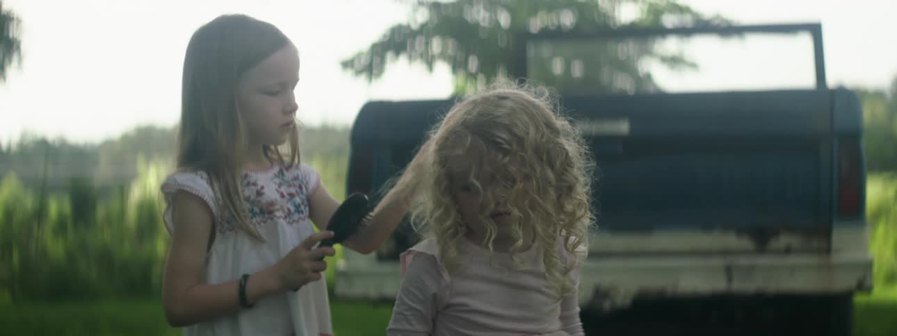 A girl brushes the hair of a smaller girl as they stand outdoors during the day