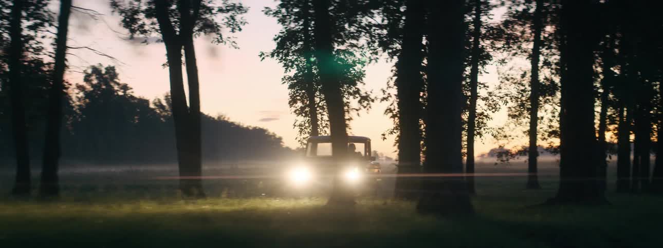 A vehicle with headlights driving on a grassy field with trees the sky and clouds in the background
