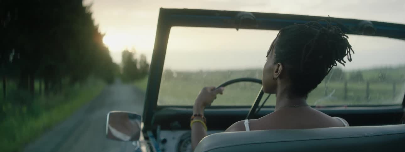 A young woman drives a vintage vehicle down a dirt road as the sun is low in the sky