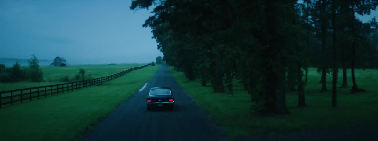 Car drives along road paralleling pasture fence with building standing in distance backed by fog and hills