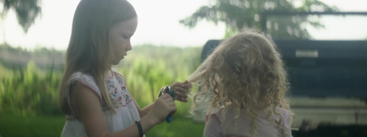 A young girl brushes the hair of her younger sister while they stand outside