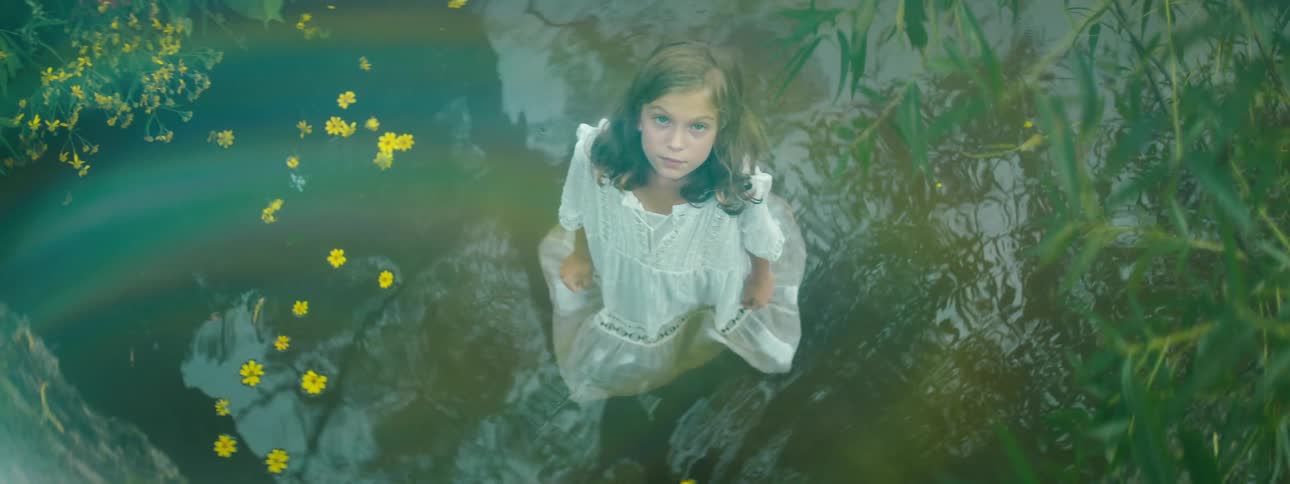 A girl in a white dress standing in a pond staring surrounded by plants and flowers