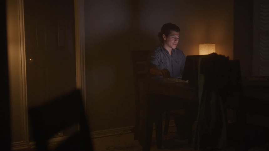 A man smokes a cigarette while using a laptop at a table in a dimly lit room