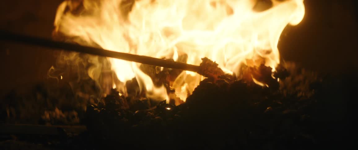 A large bonfire burns outside as a fire poker moves wood around