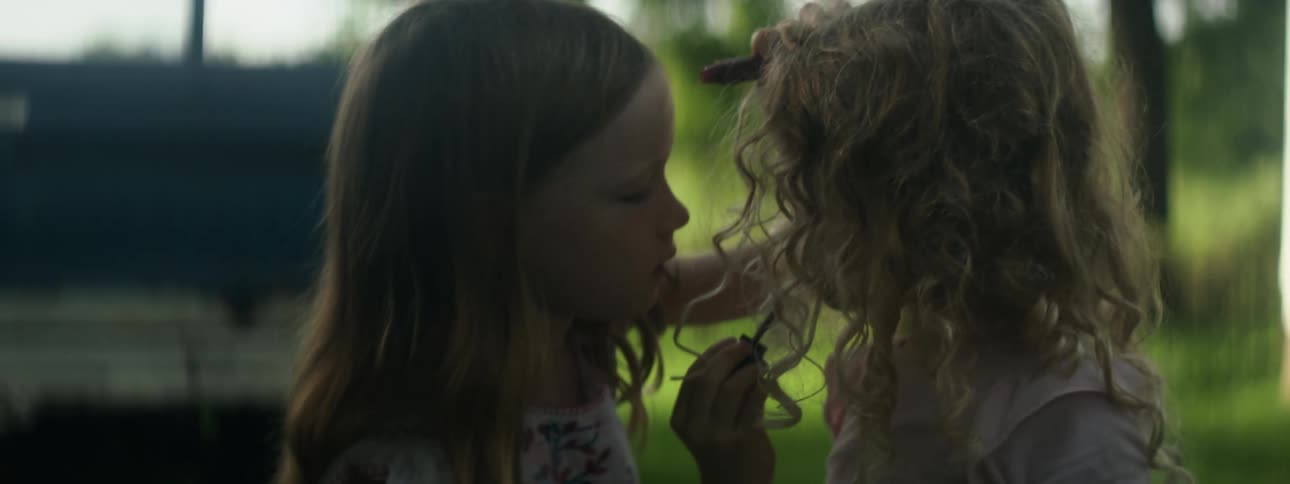 Older sister puts lipstick on younger sister as they stand outdoors between vehicle and house