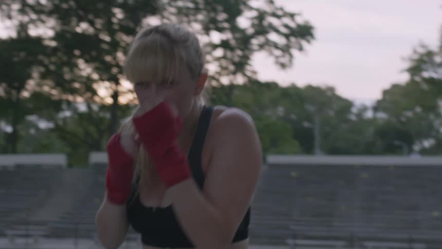 A woman practices her boxing moves in an outdoor area with bench seating in the background