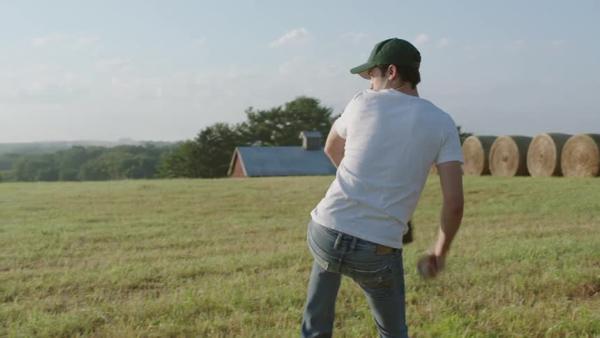 A teenage standing in an open field on a farm pitches a baseball