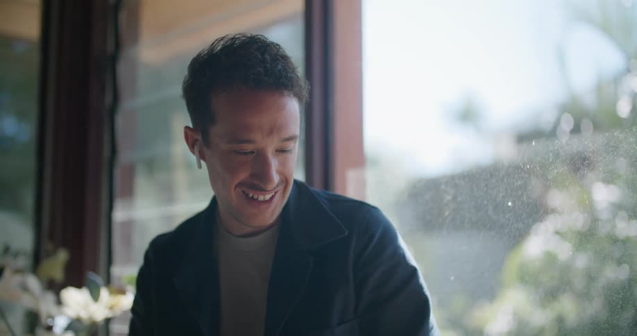 Man sits on a window ledge and smiles as he looks at a laptop