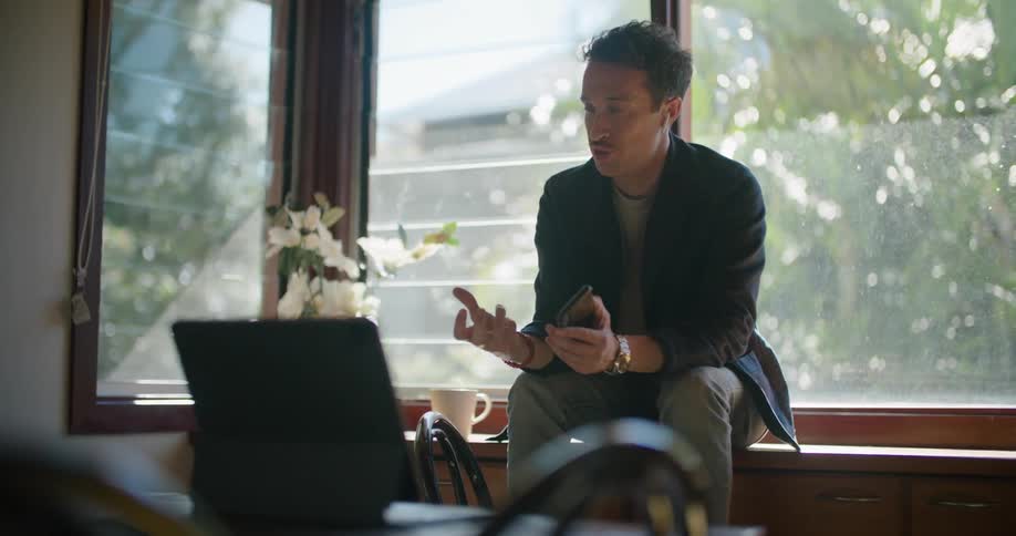 Man sits on furniture next to window holding mobile phone and gesturing while talking across from laptop sitting on dining table