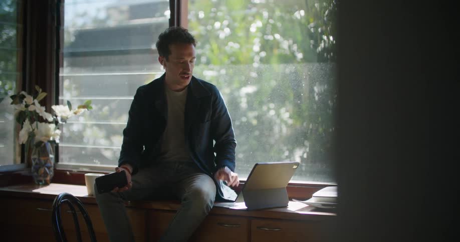 A man sits next to a window while he has a conversation with someone via his tablet computer