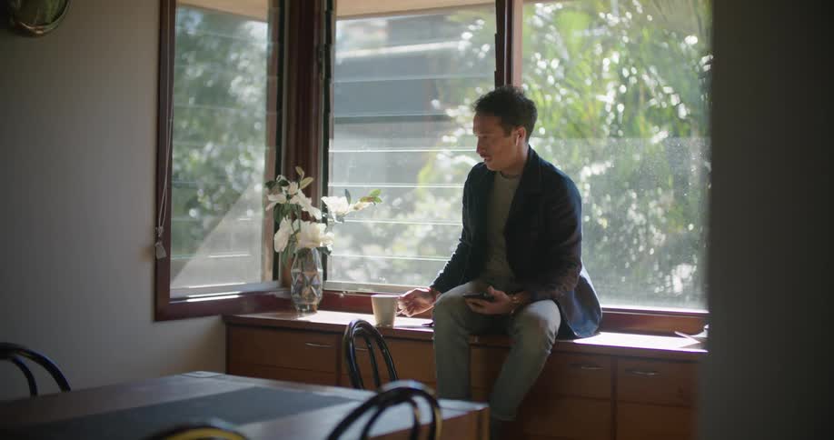 A man sits atop a cabinet located next to a large window in his kitchen