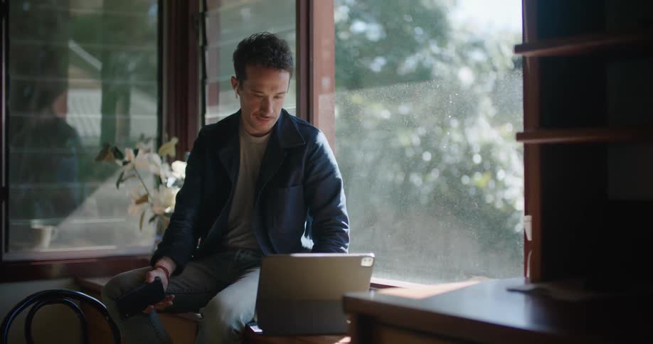 A man sitting on a counter talking on a phone and typing on a laptop