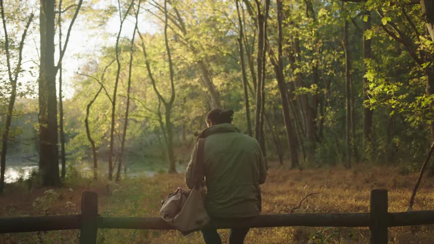 A man sits on a simple wooden fence located within a forested area