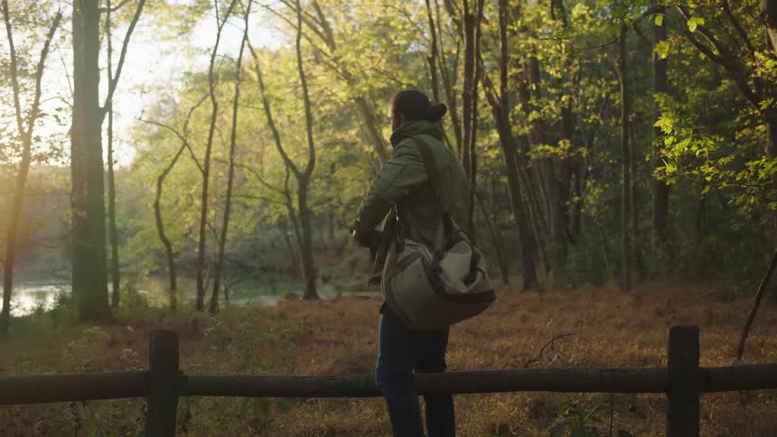 Man gets up after sitting on a wooden fence near the forest and puts a tablet in his bag and walks away