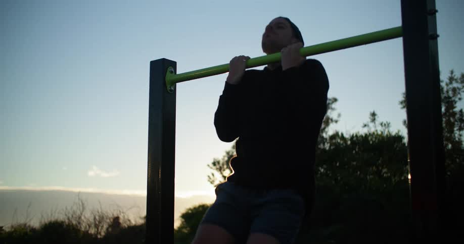 Man exercises by doing chin ups outdoors as sun sits just above the horizon behind him