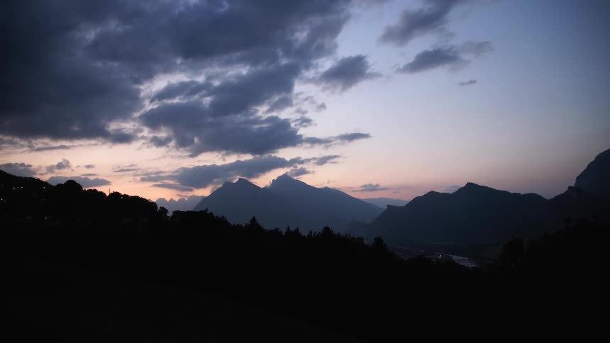 Forest trees are cast into silhouette by light from red tinted sky sitting behind misty mountains under cloudy sky