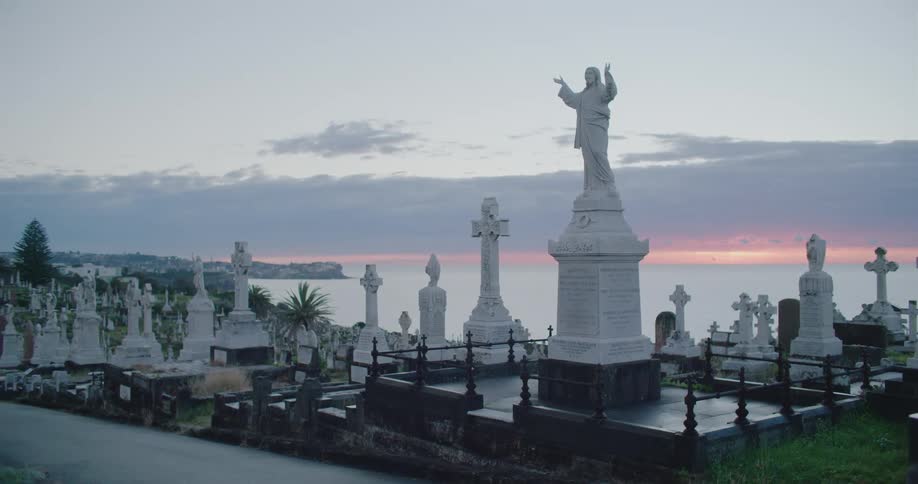 Grave monuments crowd together in graveyard with town and red horizon in distance under cloudy sky