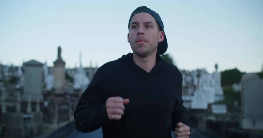 A man jogging in a cemetery with gravestones trees the sky and clouds in the background