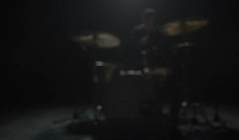A person in a room sitting playing the drums with drumsticks with a wall in the background