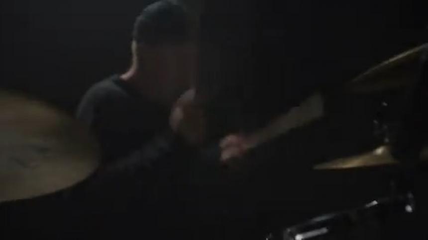 A man in a room sitting playing the drums with drumsticks with a wall in the background